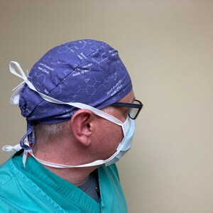 May include: A person wearing a blue surgical cap with a white pattern of chemical structures and the text "Sweet dreams are made of this". The cap has white ties that are tied at the back of the neck. The person is also wearing a white surgical mask and glasses.