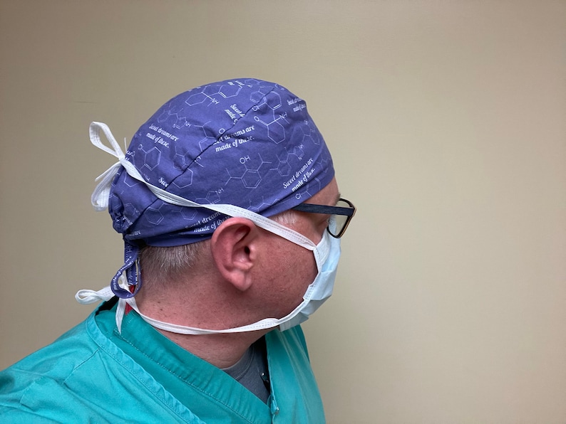 May include: A person wearing a blue surgical cap with a white pattern of chemical structures and the text "Sweet dreams are made of this". The cap has white ties that are tied at the back of the neck. The person is also wearing a white surgical mask and glasses.