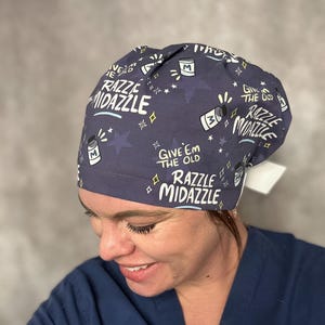 May include: A navy blue surgical scrub cap with a repeating pattern of stars, bottles, and the phrase "Give 'Em The Old Razzle Midazzle" in white. The cap is worn by a person in blue scrubs.
