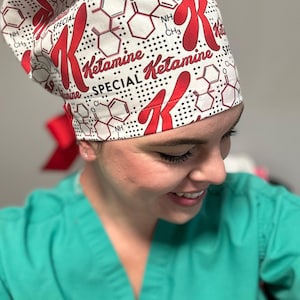K&ouml;nnte beinhalten: Eine wei&szlig;e OP-M&uuml;tze mit einem roten und schwarzen Druck. Der Druck zeigt das Wort "Ketamin" und eine chemische Formel. Die M&uuml;tze wird von einer Person mit einem gr&uuml;nen OP-Oberteil getragen.