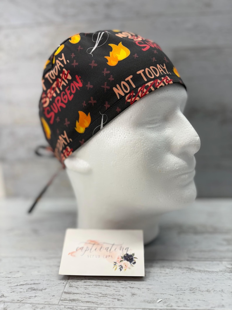 May include: Black scrub hat with a repeating pattern of flames, crosses, and the text "Not Today Satan Surgeon".