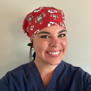 May include: Red surgical scrub cap with a repeating pattern of a cartoon bear and the letter "K" in white. The cap is worn by a person wearing a blue scrub top.