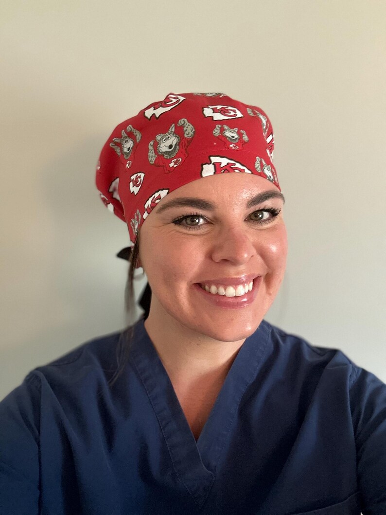 May include: Red surgical scrub cap with a repeating pattern of a cartoon bear and the letter "K" in white. The cap is worn by a person wearing a blue scrub top.