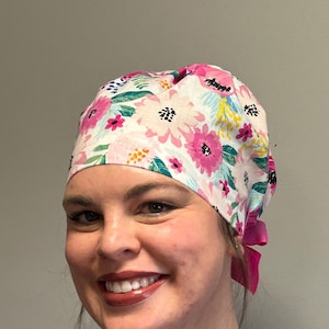 May include: A floral patterned scrub cap featuring pink, white, and green flowers on a white background. The cap has a pink ribbon tied at the back. The cap is worn by a person.