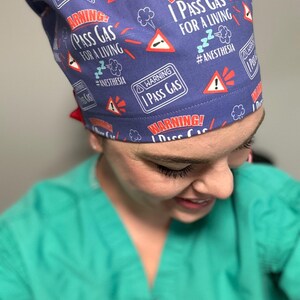 May include: A purple scrub hat with a repeating pattern of red warning signs that say "Warning! I Pass Gas For A Living #Anesthesia".