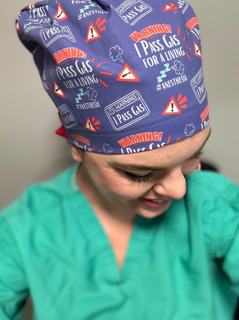 May include: A purple scrub hat with a repeating pattern of red warning signs that say "Warning! I Pass Gas For A Living #Anesthesia".