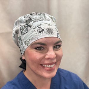 May include: A white surgical scrub cap with a black and white print featuring medical-themed graphics and the text "THE TORTURED ANESTHESIA DEPARTMENT" and "BETAMINE". The cap is tied in the back with a black ribbon.