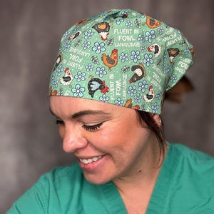May include: A scrub cap in a light green fabric featuring a repeating pattern of chickens, blue flowers, and the text "FLUENT IN FOWL LANGUAGE" printed in white. The cap has a fitted crown and is tied at the back with fabric ties.