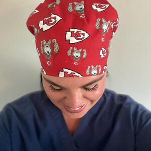 May include: Red surgical cap featuring a repeating pattern of a cartoon wolf in a football uniform and the Kansas City Chiefs logo. The cap is made of a lightweight fabric and is designed to be worn in a medical setting.
