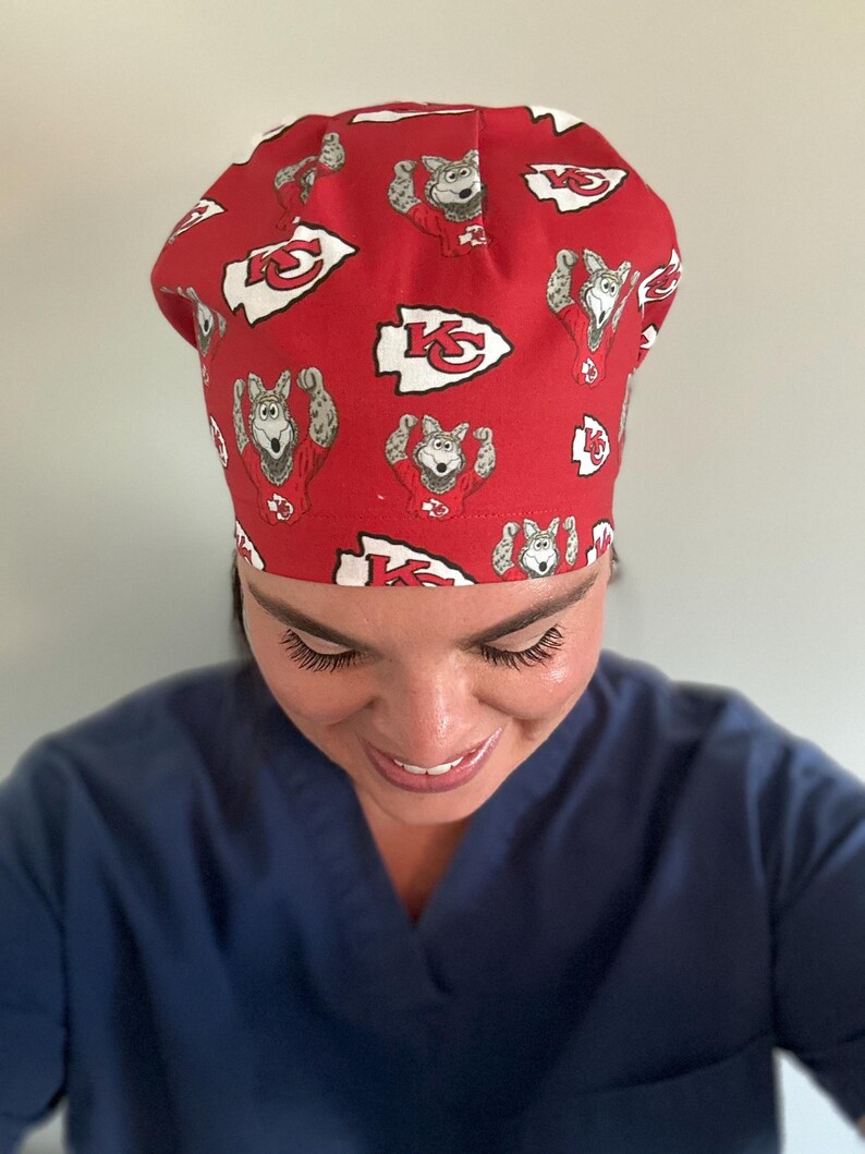 May include: Red surgical cap featuring a repeating pattern of a cartoon wolf in a football uniform and the Kansas City Chiefs logo. The cap is made of a lightweight fabric and is designed to be worn in a medical setting.