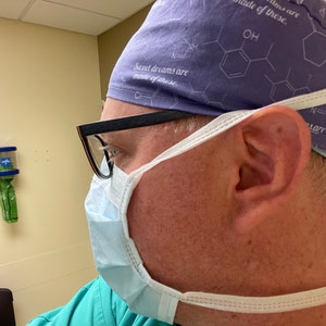 May include: A person wearing a purple surgical cap with the text "Sweet dreams are made of these." and a white surgical mask. The person is wearing glasses and a green shirt.