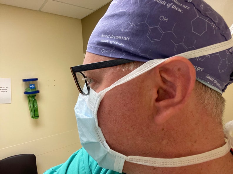 May include: A person wearing a purple surgical cap with the text "Sweet dreams are made of these." and a white surgical mask. The person is wearing glasses and a green shirt.