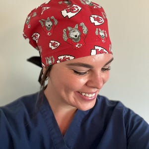 May include: Red scrub cap featuring a repeating pattern of a cartoon wolf in a football jersey and the Kansas City Chiefs logo. The cap is tied in the back with a black ribbon. The person in the image is wearing a blue scrub top.