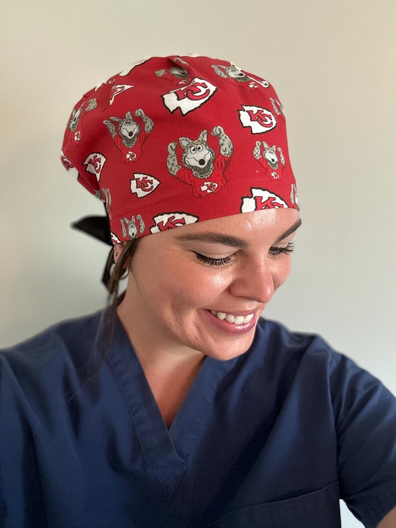 May include: Red scrub cap featuring a repeating pattern of a cartoon wolf in a football jersey and the Kansas City Chiefs logo. The cap is tied in the back with a black ribbon. The person in the image is wearing a blue scrub top.