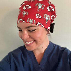 May include: Red surgical scrub cap with a repeating pattern of a cartoon wolf in a football jersey and the Kansas City Chiefs logo. The cap is tied in the back with a black bow.