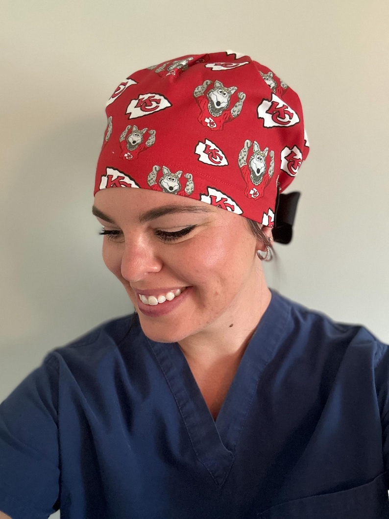 May include: Red surgical scrub cap with a repeating pattern of a cartoon wolf in a football jersey and the Kansas City Chiefs logo. The cap is tied in the back with a black bow.
