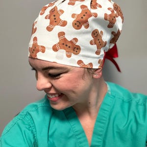 May include: A white scrub hat with a gingerbread man pattern and a red tie. The hat is worn by a person with a green scrub top.