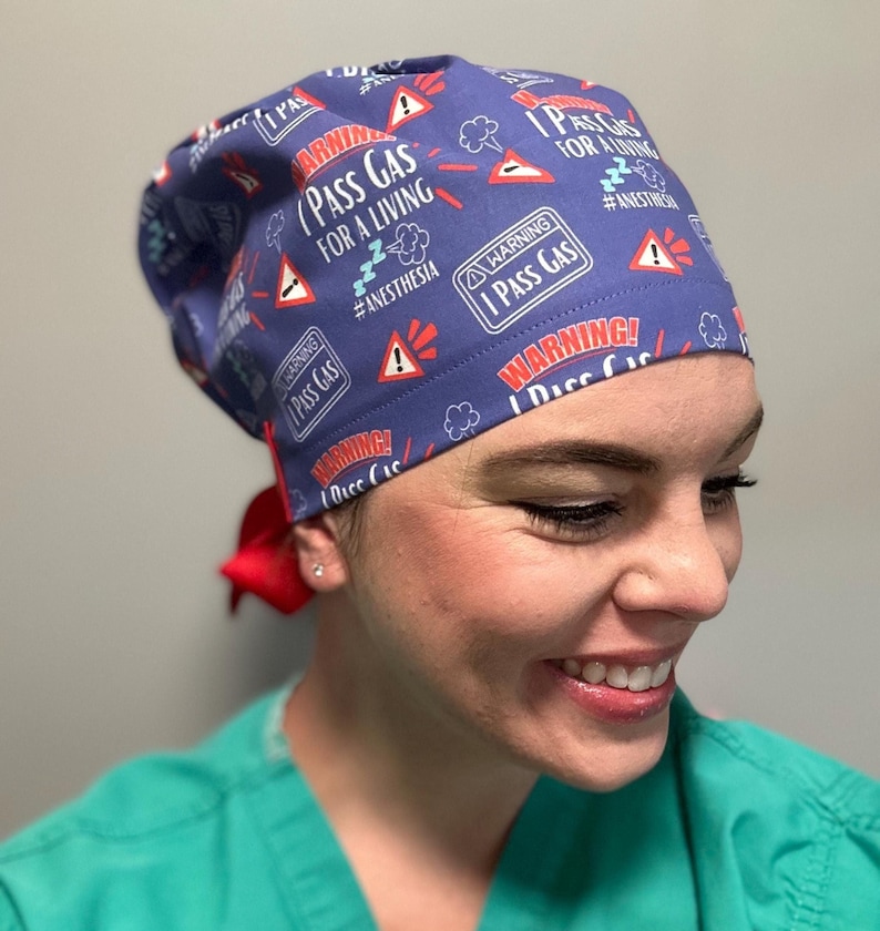May include: A blue scrub hat with a red tie, featuring a repeated pattern of white warning signs that say "Warning! I Pass Gas for a Living #Anesthesia".