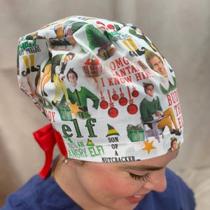 May include: White surgical scrub cap with a festive "Elf" movie theme. The cap features images of the movie's characters, Christmas ornaments, and text like "OMG Santa! I know him!" and "Son of a Nutcracker."