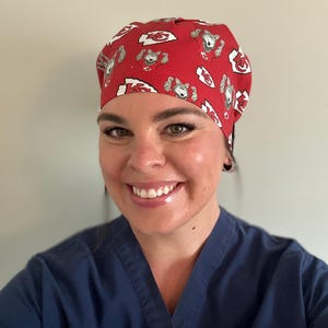 May include: Red scrub cap with a repeating pattern of cartoon wolf characters and the Kansas City Chiefs logo. The cap is made of a soft fabric and is designed to be worn in a medical setting.