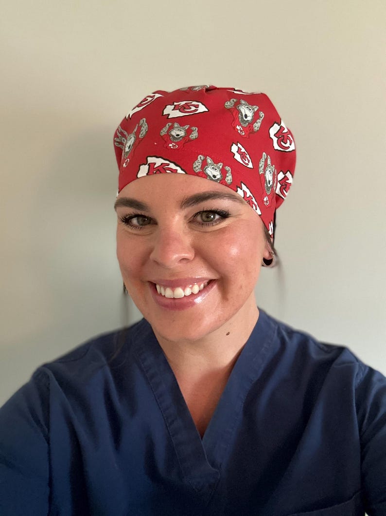 May include: Red scrub cap with a repeating pattern of cartoon wolf characters and the Kansas City Chiefs logo. The cap is made of a soft fabric and is designed to be worn in a medical setting.
