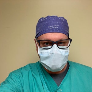 May include: A man wearing a purple surgical scrub cap with the text "Sweet dreams are made of these." He is also wearing a blue surgical mask and green scrubs.