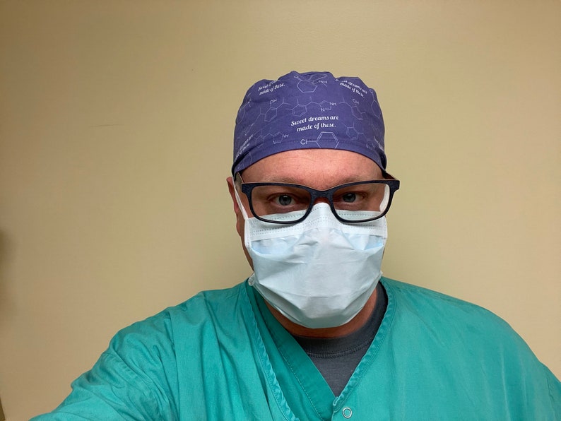 May include: A man wearing a purple surgical scrub cap with the text "Sweet dreams are made of these." He is also wearing a blue surgical mask and green scrubs.