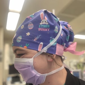 May include: Purple surgical scrub hat with a pink tie. The hat has a pattern of Easter eggs, bunnies, carrots, and syringes. The text "Ether Bunny" is printed on the hat.