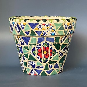 May include: A decorative mosaic flower pot with a vibrant design. The pot features a mosaic pattern of green, blue, yellow, and red tiles. The rim is lined with small green tiles. The pot is suitable for indoor or outdoor use.