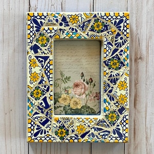 May include: Rectangular photo frame with a mosaic tile border. The frame's border features a pattern of blue, yellow, and white tiles. The frame holds a picture of roses and script. The frame is approximately 6 inches tall and 4 inches wide.