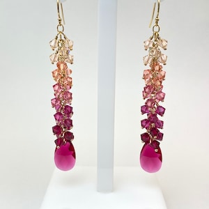 May include: A pair of long, gold dangle earrings with a gradient of pink and red crystals. The earrings are made with small, faceted crystals that are strung together in a cascading design. The bottom of each earring features a large, teardrop-shaped crystal.