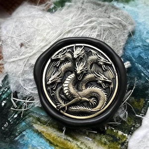 May include: A circular wax seal featuring a detailed metallic gold relief of a three-headed dragon with outstretched wings. The dragon is centred within a smooth, thick black wax border, showcasing intricate scales and sharp facial features.