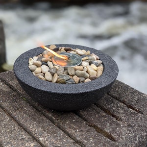 May include: A black fire pit with a small flame burning in the center. The fire pit is filled with small gray stones and is sitting on a wooden surface.