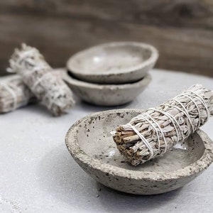 May include: A close-up shot of a white sage smudge stick resting in a small, gray, concrete bowl. Several other bowls and smudge sticks are in the background. The sage is tied with white string. The bowls have a speckled texture.