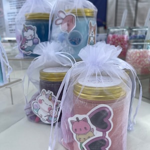Puede incluir: Tres frascos de vidrio llenos de slime de colores, cada uno en una bolsa de organza blanca con una pegatina de un personaje de dibujos animados lindo. Los colores del slime son azul, morado y rosa.