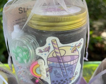 Könnte beinhalten: Eine lila und weiße Geschenktüte gefüllt mit einem durchsichtigen Glas mit goldenem Deckel, einem kleinen durchsichtigen Glas mit grünem und weißem Design, einem Regenbogen-Anhänger und einer kleinen Flasche Flüssigkeit. Das Glas hat einen Aufkleber mit einem lila Boba-Tee mit einem Strohhalm und Fischen, die darin schwimmen.