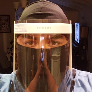 May include: A person wearing a black face shield with the text "Face Shield" printed on the top. The person is also wearing a black balaclava and a blue shirt.