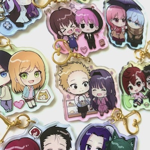 May include: A collection of clear acrylic keychains featuring various pairs of illustrated anime-style characters. Each keychain includes a gold-colored heart-shaped clasp and displays colorful, chibi-proportioned figures in diverse outfits and settings.