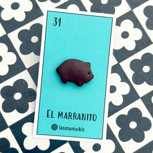 May include: A dark brown pig-shaped pin on a teal card with the number 31. The card reads "EL MARRANITO" and has the Instagram handle "@lasmamukis". The background is a black and white floral pattern.