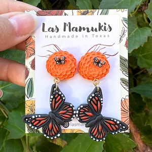 May include: A pair of handmade earrings featuring orange flower-shaped studs with small black butterflies. Below, large black butterfly pendants with orange and white accents dangle from gold-colored hooks. The earrings are displayed on a card that reads "Las Mamukis Handmade in Texas."