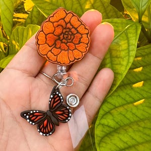 May include: A retractable badge reel featuring an orange flower and a butterfly with black, red, and white details. The badge reel has a clear plastic strap and a metal clip for attaching to clothing.