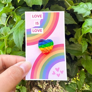 May include: A rainbow heart-shaped pin with the text "LOVE IS LOVE" on a pink card with rainbow stripes and pink hearts.