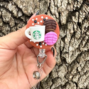 May include: A brown and white polka dot badge reel with a white coffee cup featuring the Starbucks logo, a brown and white concha, and a pink and green concha.
