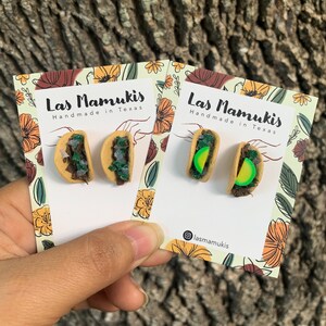 May include: Two pairs of taco-shaped earrings on a white card with the text "Las Mamukis Handmade in Texas" and the Instagram handle "@lasmamukis". The earrings are made of polymer clay and feature a green lettuce, brown meat, and a yellow lime.