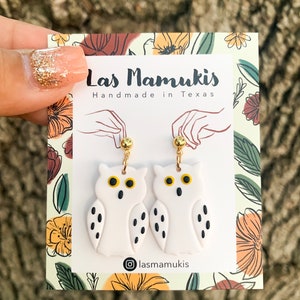 May include: White owl shaped earrings with black dots and gold accents. The earrings are on a card with the text "Las Mamukis Handmade in Texas" and the Instagram handle "@lasmamukis".