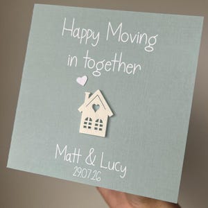 Może przedstawiać: Jasnoniebieska kartka z napisem "Happy Moving in together" oraz imionami "Matt & Lucy" z datą. Do kartki przymocowany jest mały drewniany domek z wyciętym sercem.