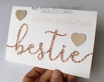 Puede incluir: Una tarjeta blanca con las palabras "Happy Birthday bestie" en letras de purpurina dorada rosa. La tarjeta tiene dos pequeños corazones de purpurina dorada rosa.