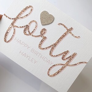 May include: A white card with the word "forty" written in gold glitter script. The word "Happy Birthday" is written in pink script below the word "forty". The name "Hayley" is written in pink script below the word "Birthday". A small silver glitter heart is adhered to the top of the card.