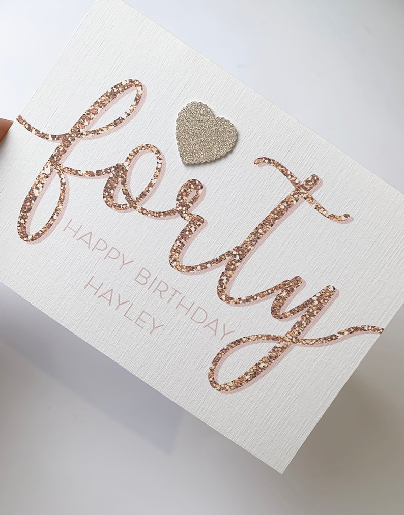 May include: A white card with the word "forty" written in gold glitter script. The word "Happy Birthday" is written in pink script below the word "forty". The name "Hayley" is written in pink script below the word "Birthday". A small silver glitter heart is adhered to the top of the card.