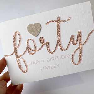 May include: A white card with the word "forty" written in gold glitter. The word "happy birthday" is written below the word "forty" in black text. The name "Hayley" is written below the word "happy birthday" in black text. A small gold glitter heart is in the upper left corner of the card.